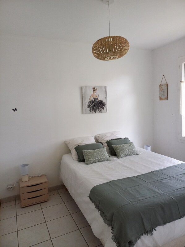 Un dormitorio con cama, almohadas, una lámpara colgante, una obra de arte en la pared y un pequeño cofre de madera.