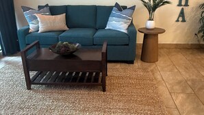 A teal sofa with patterned cushions, a wooden coffee table with a bowl, a wall-mounted paddle decoration, and a potted plant. - **Newly Renovated** Ground Floor Condo In Paradise (Kihei)
