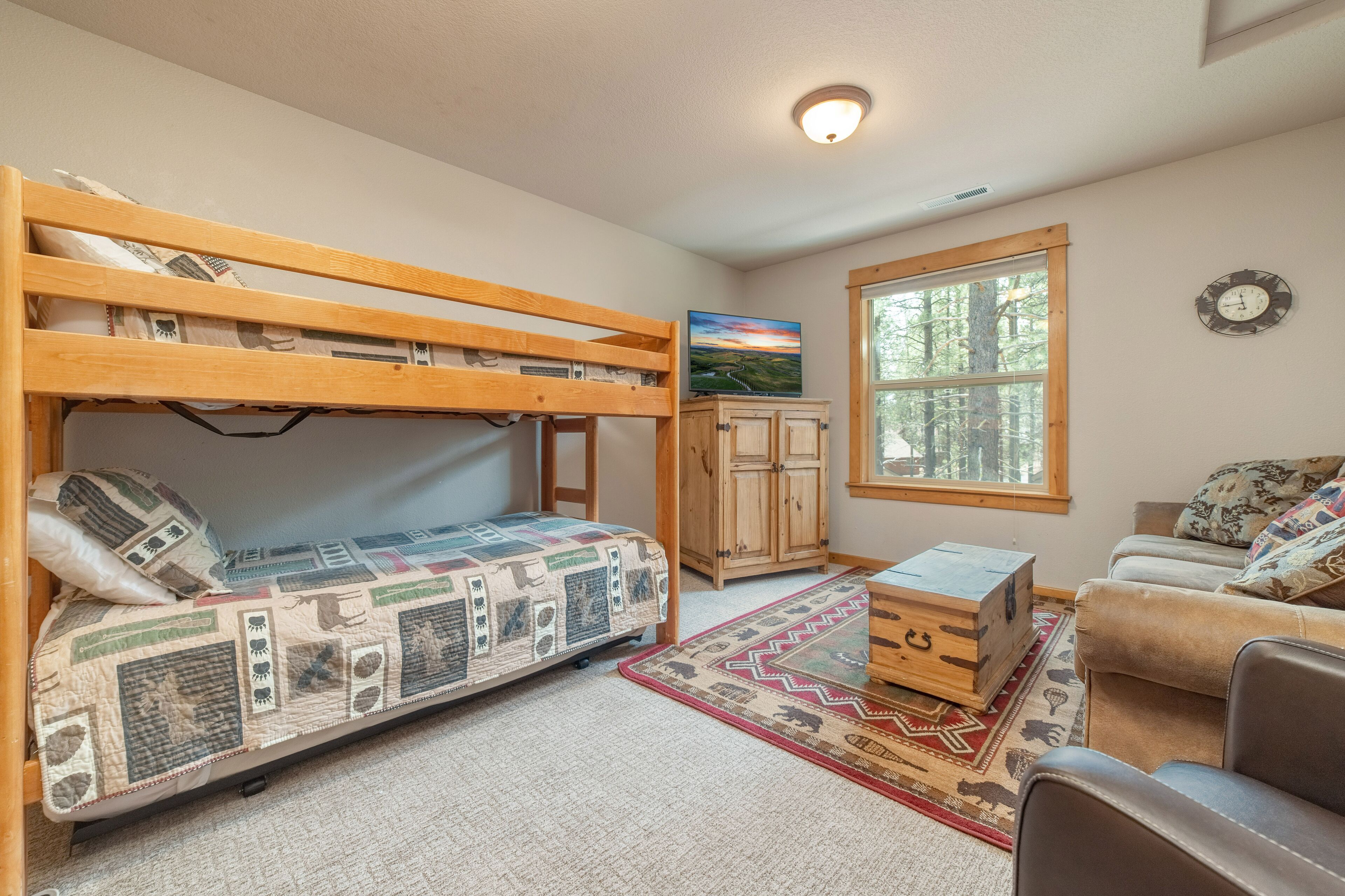 A bunk bed with a quilt, a wooden cabinet, a television, a window with a view of trees, and a couch.