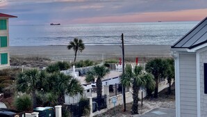 A coastal view with a beach, palm trees, and a building with a white fence. - 30 Steps to the Beach! Luxury Condo with Large wrap-around porch! (Tybee Island)
