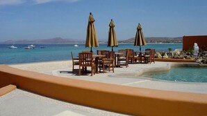 Un espace aménagé au bord de la piscine, avec des sièges et des parasols, offrant une vue sur une mer d’un bleu limpide où naviguent des bateaux.