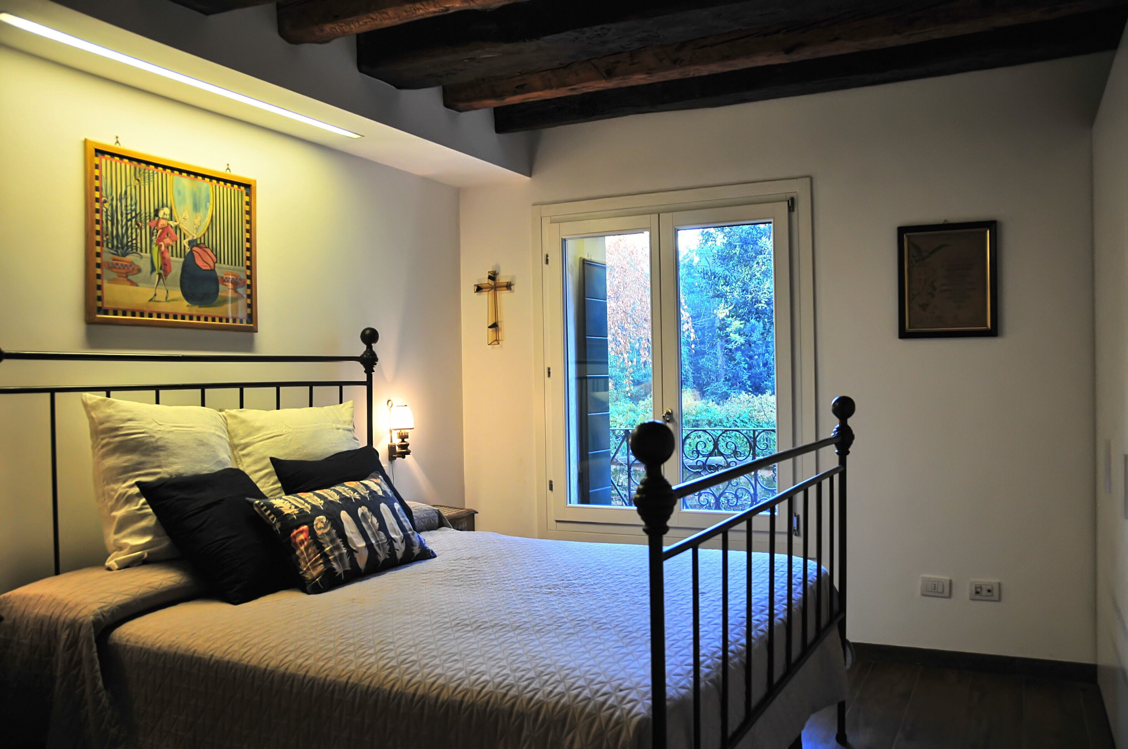 A bedroom with a bed, a nightstand, a wall-mounted cross, and a window with a view of greenery.