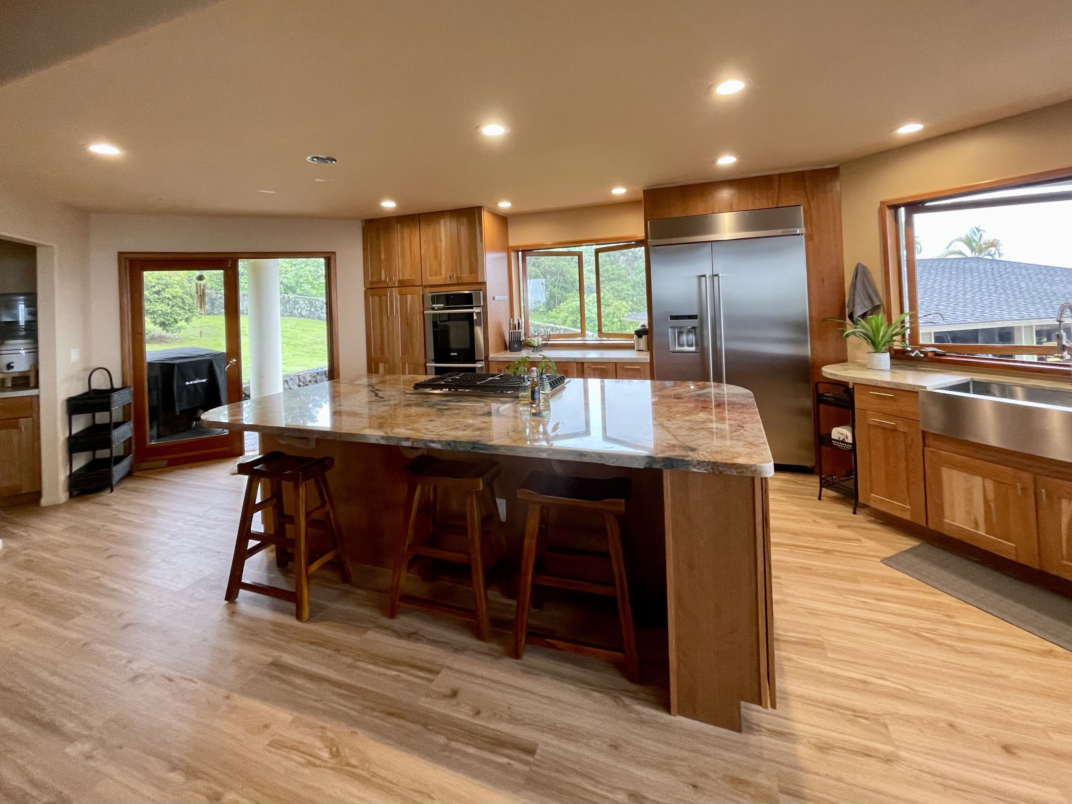 Une cuisine moderne dotée d’un grand îlot central, de meubles en bois, d’appareils électroménagers en acier inoxydable et offrant une vue sur l’extérieur.