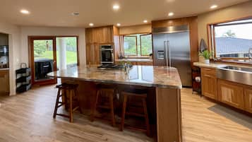 Une cuisine moderne dotée d’un grand îlot central, de meubles en bois, d’appareils électroménagers en acier inoxydable et offrant une vue sur l’extérieur.