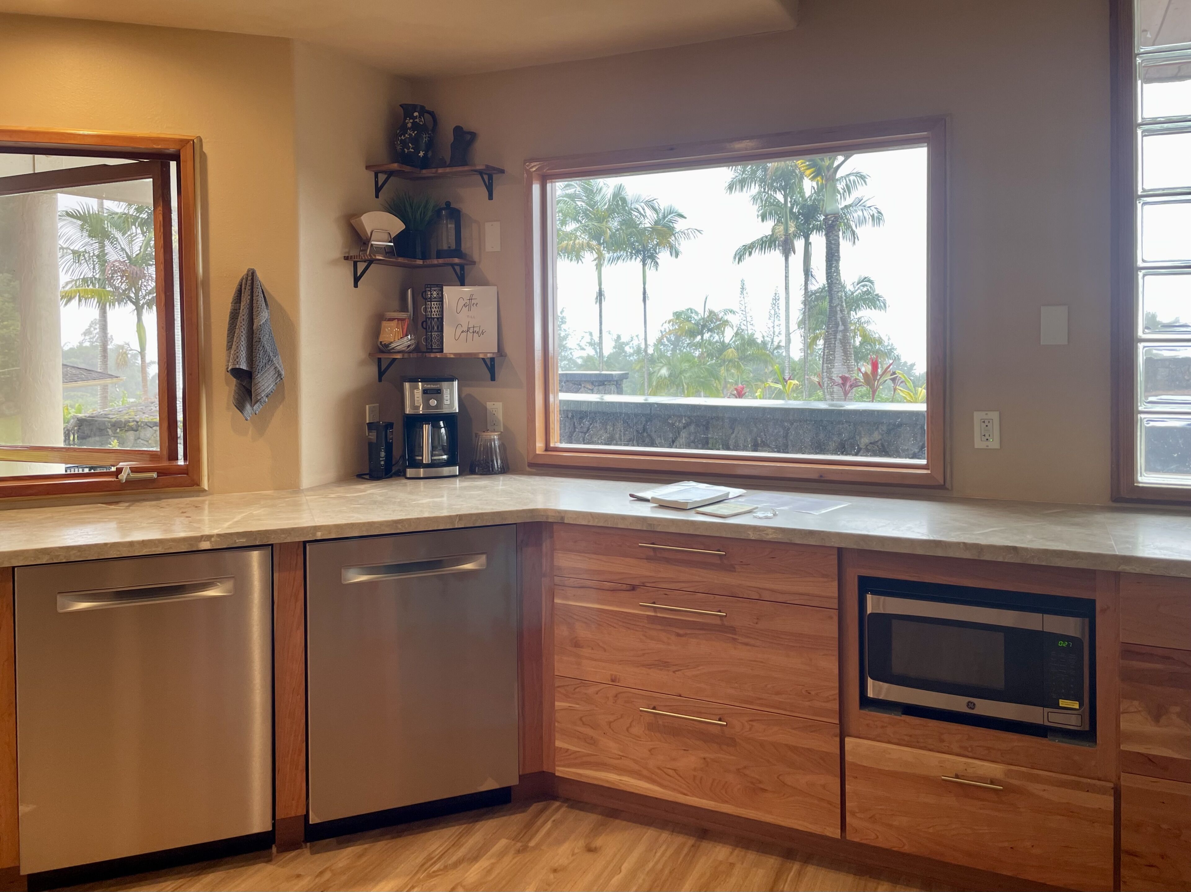 Una cocina moderna con lavavajillas integrado, armarios de madera, estación de café y un amplio ventanal con vistas a palmeras.