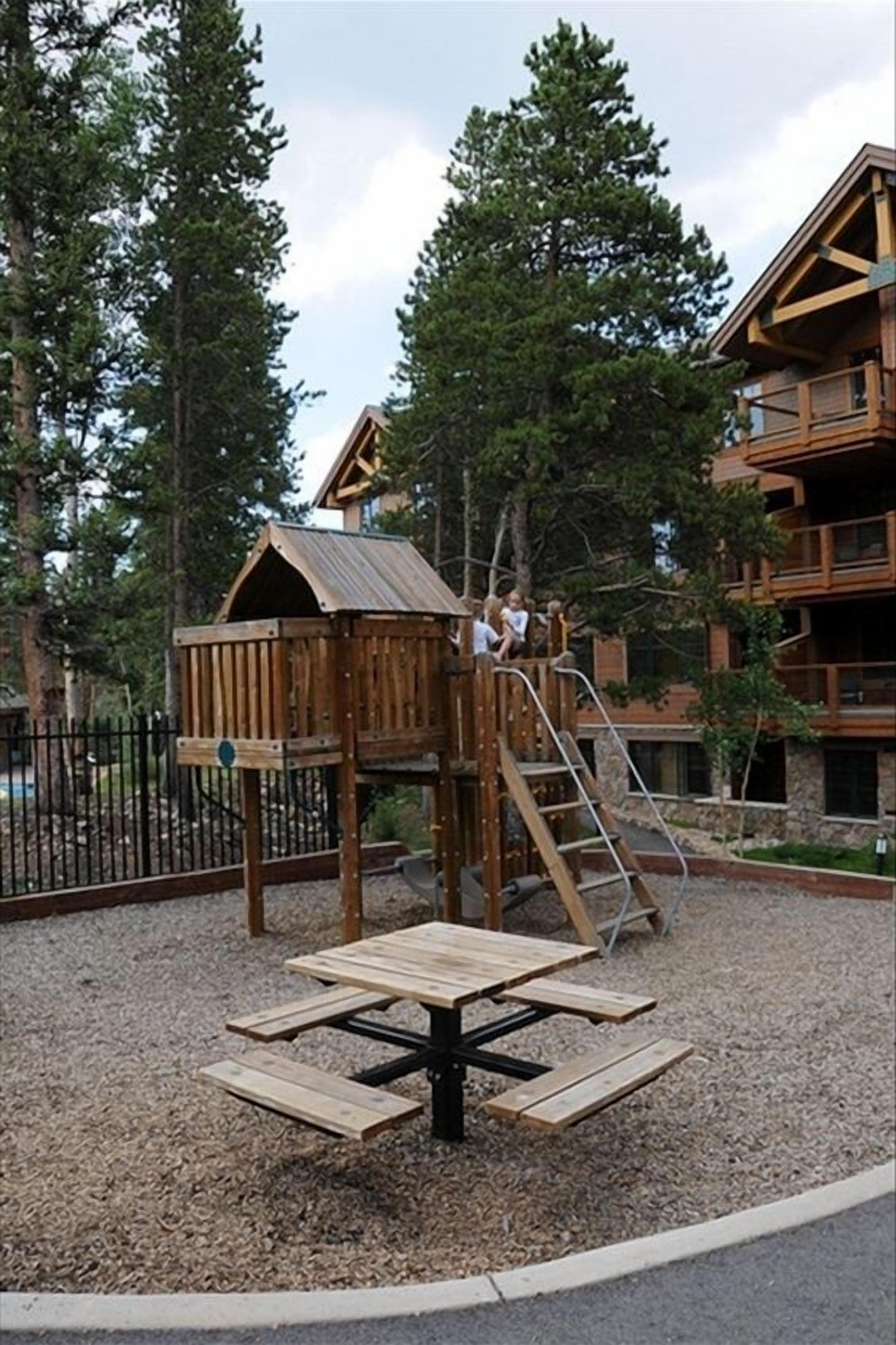 A wooden play structure with a slide and a picnic table in a wooded area with a building in the background.