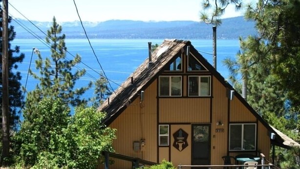 A wooden cabin with a steep gabled roof, a front porch, and a view of a lake and mountains in the background.