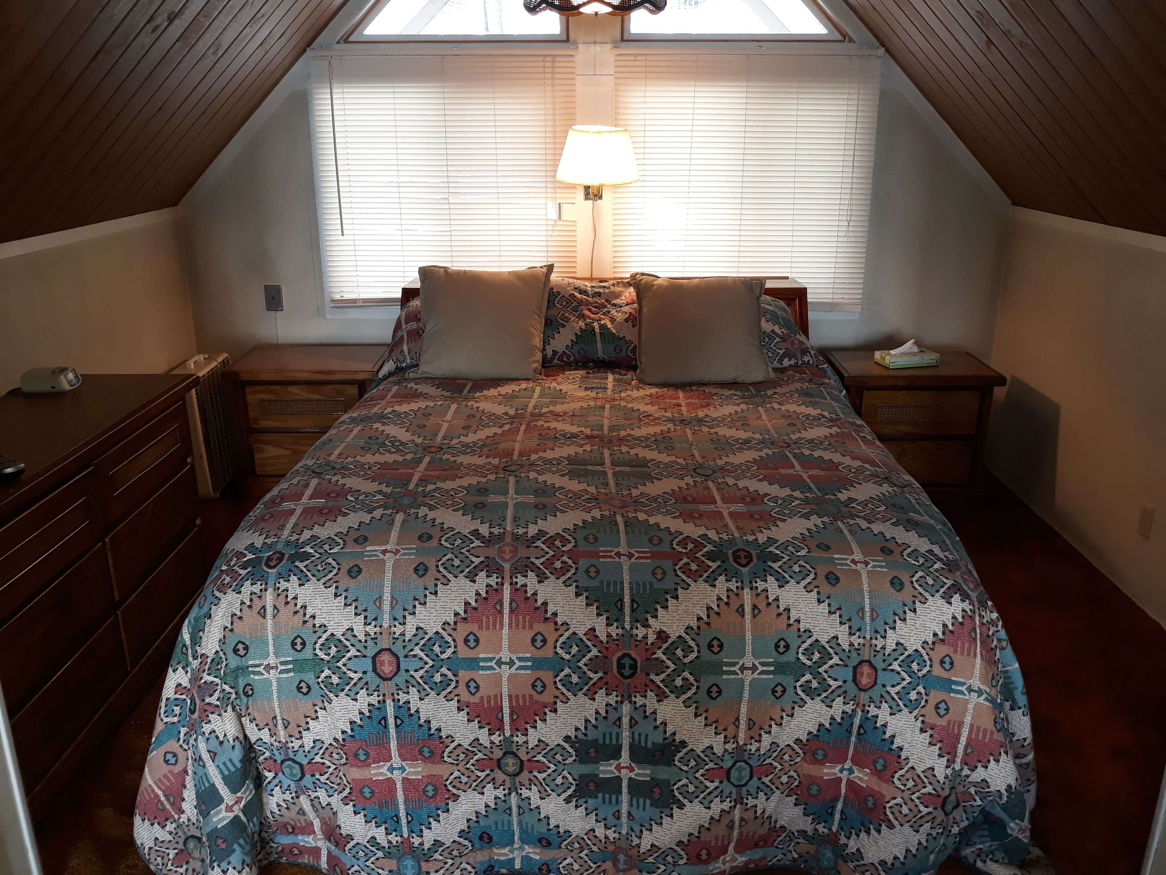 A bedroom with a sloped ceiling, a bed with patterned bedding, two bedside lamps, and a window with blinds.
