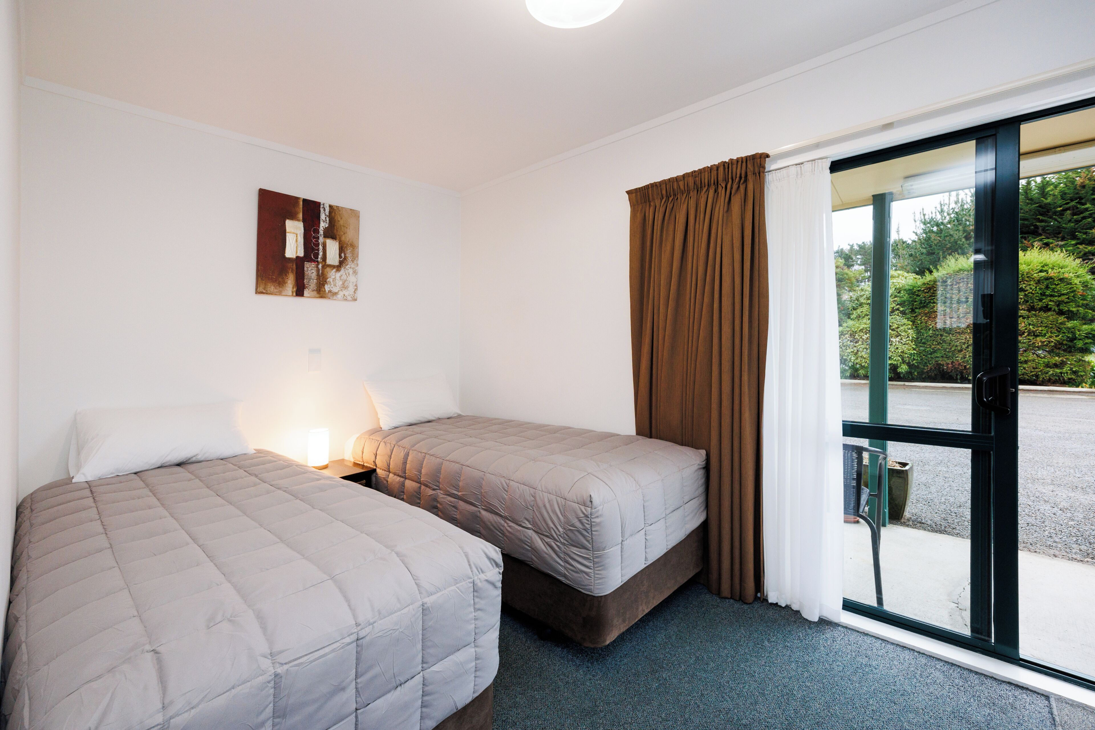 A room with two beds, a window with curtains, a wall-mounted artwork, and a view of the outside.