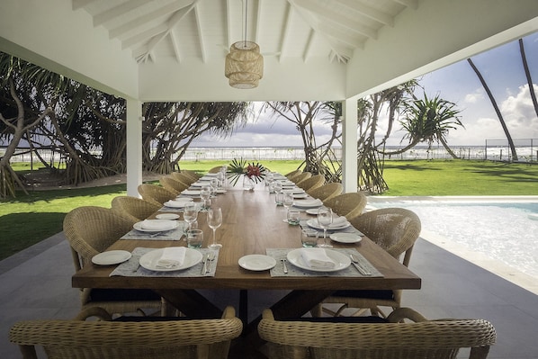 Ein langer Esstisch, gedeckt für eine Mahlzeit, mit Korbstühlen, mit Blick auf einen Pool und eine Rasenfläche mit Bäumen.