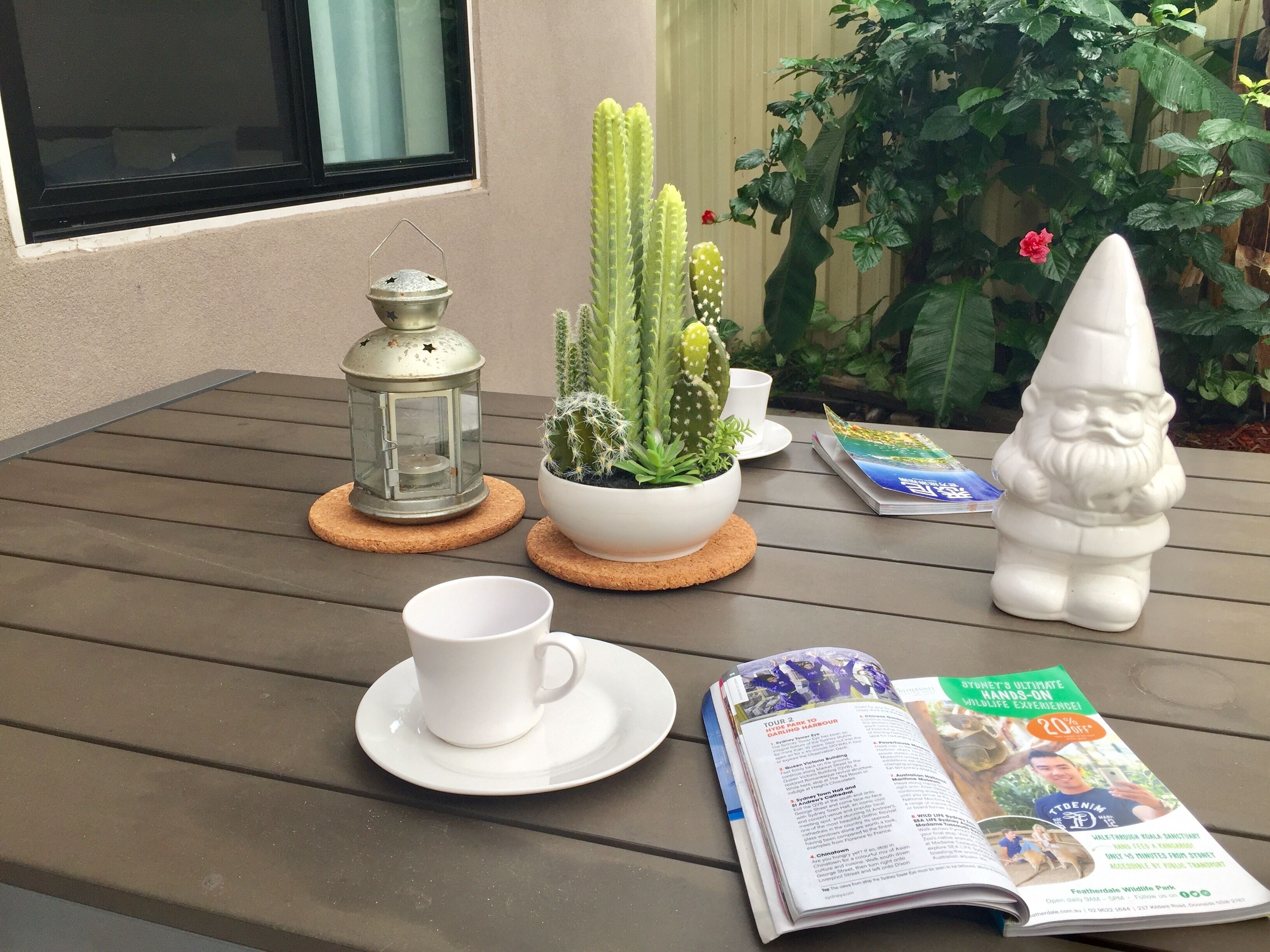 A table with a white cup, saucer, a metal lantern, a potted cactus, a gnome statue, and a magazine.