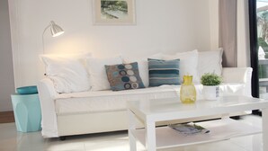 Ein weißes Sofa mit gemusterten Kissen, ein weißer Couchtisch, ein gerahmtes Landschaftsbild und ein kleiner Topfpflanze.