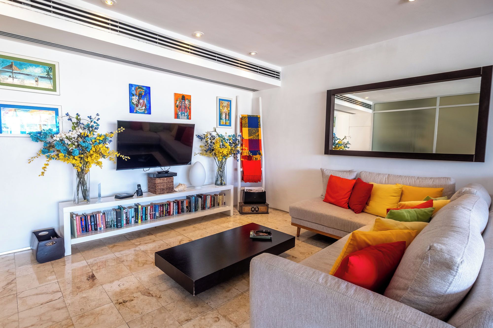 Una sala de estar moderna con un televisor de pantalla plana, una estantería, un sofá con cojines de colores y un espejo grande.