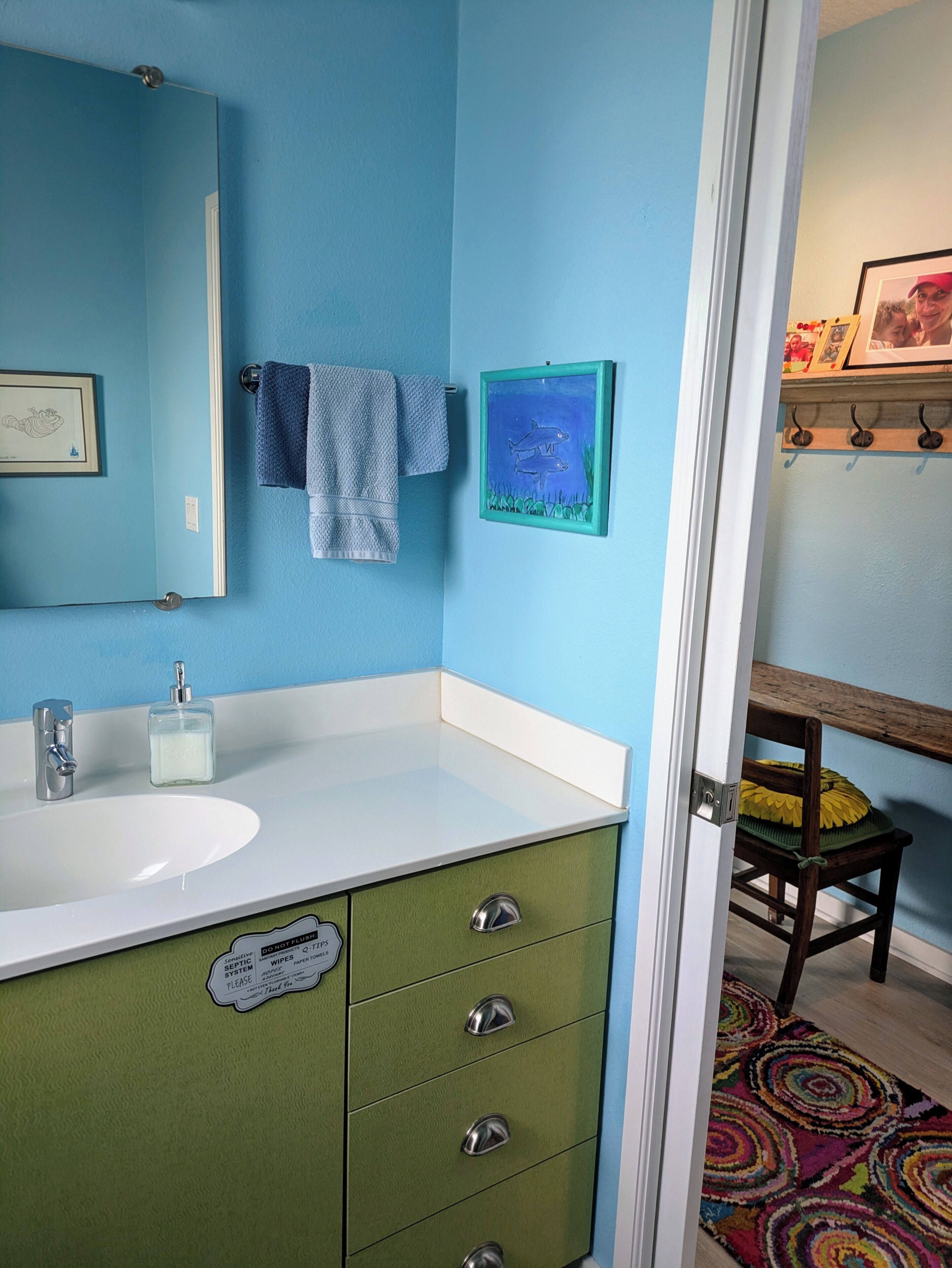 Un bagno con un lavandino bianco, mobili verdi, una parete blu, un quadro di uno squalo incorniciato e una sedia di legno con cuscino giallo.