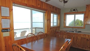 A wooden cabin with a dining table and chairs, a kitchen with wooden cabinets, and large windows offering a view of the water.