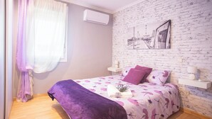 A bedroom with a brick-patterned wall, a bed with a purple and white floral bedspread, a purple throw blanket, and a small bedside table with a lamp. - Casa Melchor for 9 people (Castejón)