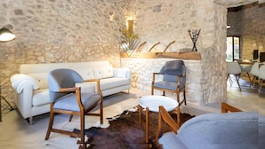 A stone-walled living room with a white sofa, two wooden chairs, a small table, and a cowhide rug.
