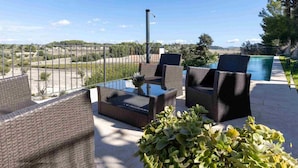 An outdoor patio with wicker furniture, a glass table, and a view of a pool and landscape.