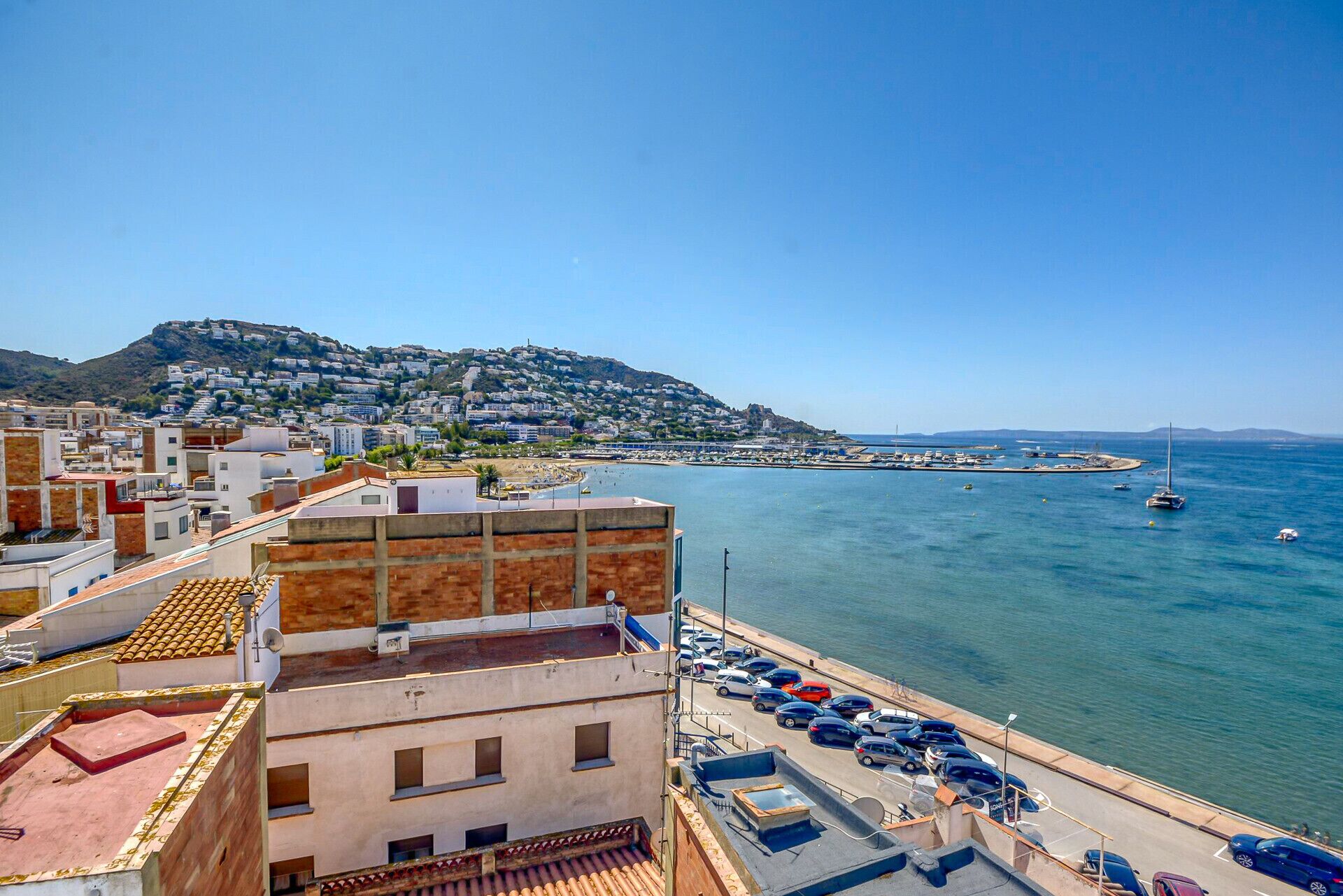 Un paysage urbain côtier avec une colline imposante, un port de plaisance avec des bateaux et un parking avec des voitures.