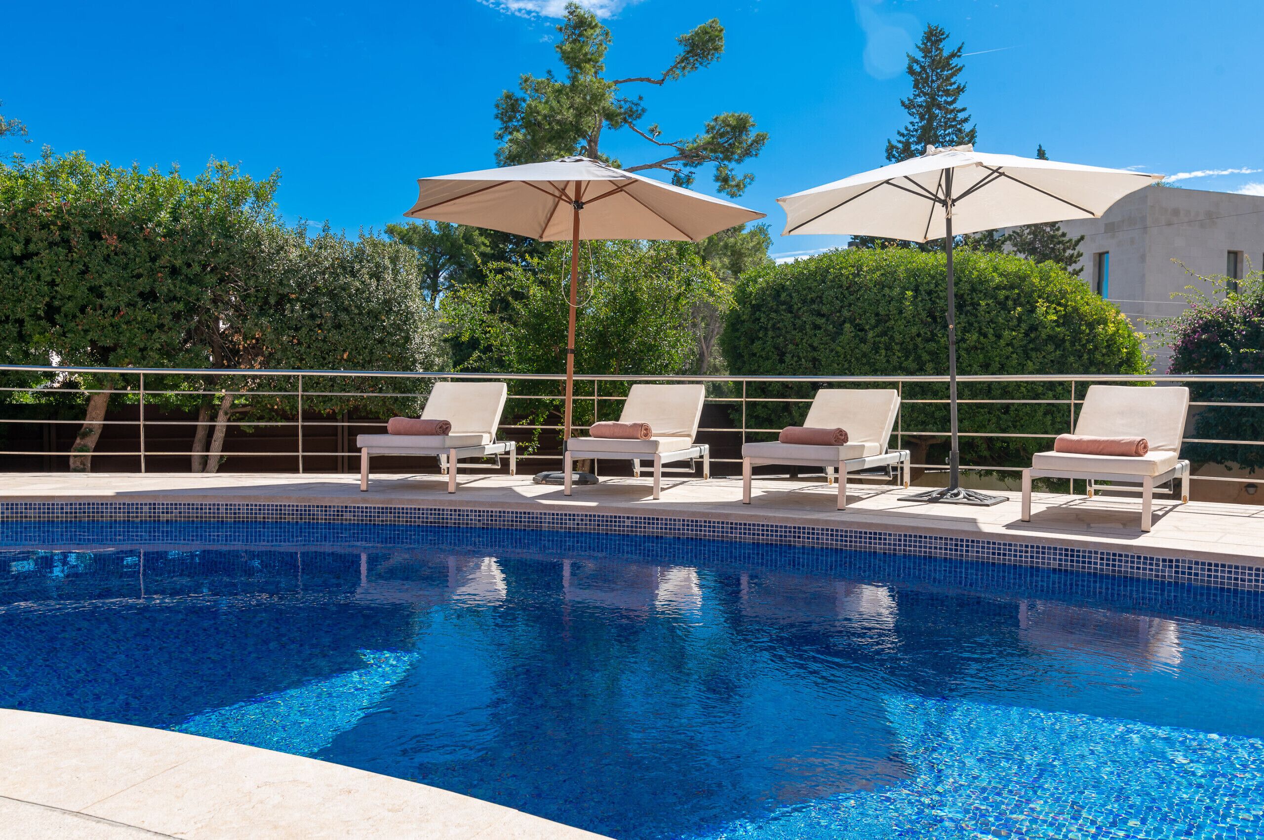 Una piscina con sillas de descanso y sombrillas, rodeada de vegetación y un cielo azul despejado.