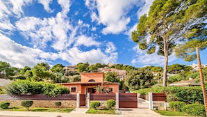 Una villa de estilo mediterráneo con un muro de piedra, techo de tejas y un jardín con arbustos y un árbol pequeño.
