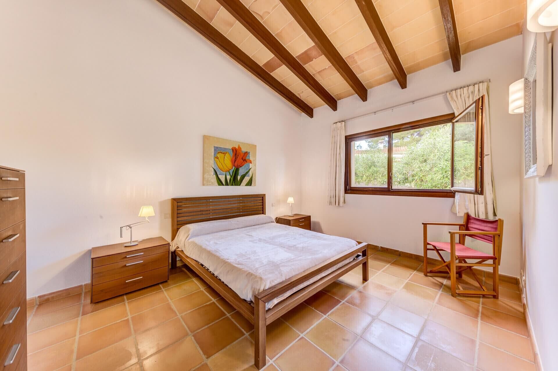 Un dormitorio con cama de madera, mesita de noche, una silla, ventana con cortinas y un cuadro en la pared.