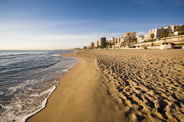 Uma praia arenosa com pegadas, céu claro e edifícios ao fundo.