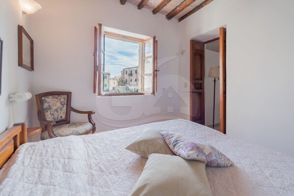 Une chambre à coucher avec un lit, une chaise, une fenêtre donnant sur des immeubles et une porte.