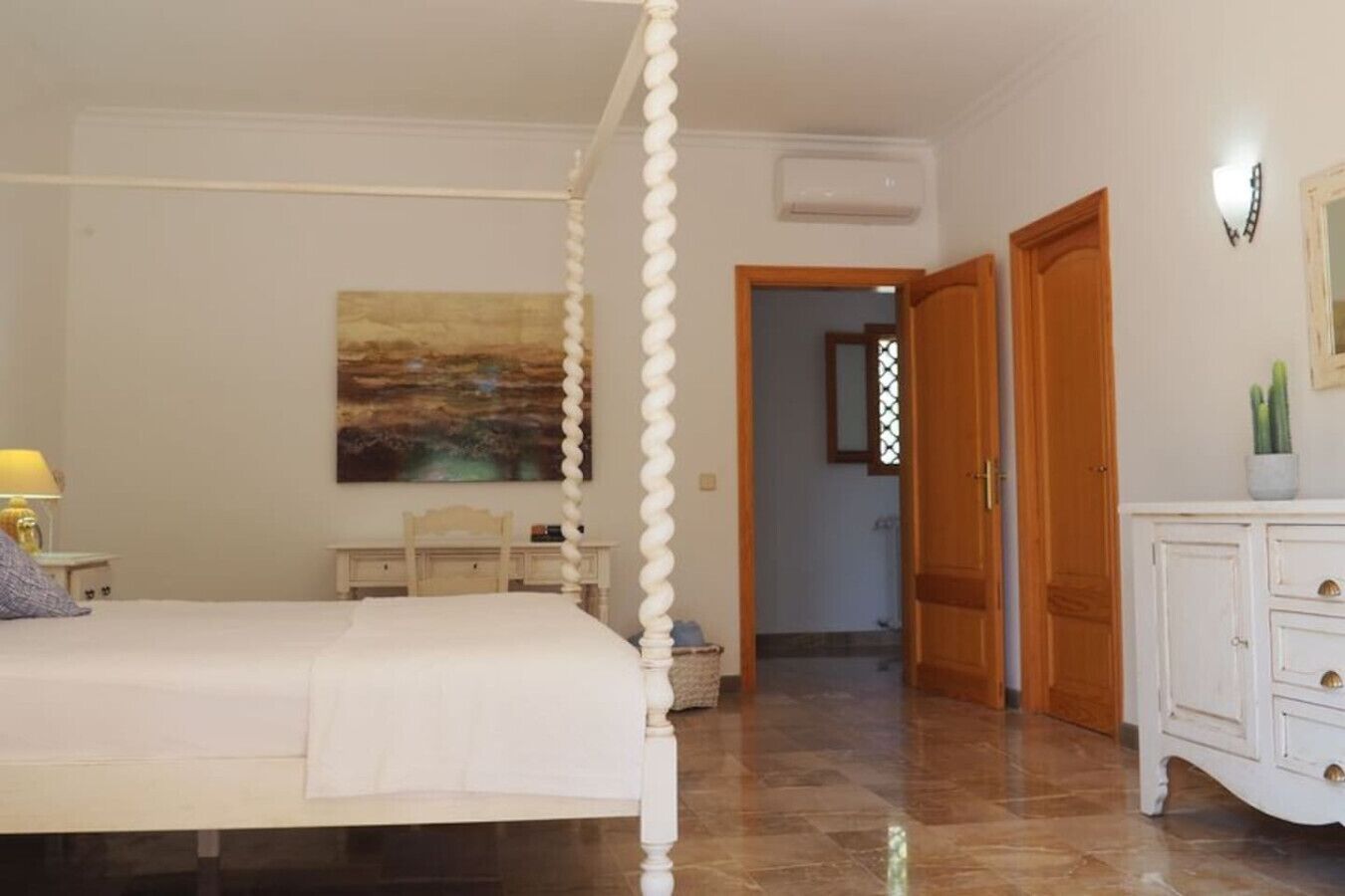 A bedroom with a four-poster bed, a bedside table, a lamp, a painting on the wall, and a wardrobe.
