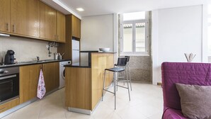 Une cuisine compacte avec des meubles en bois, une machine à café et un lave-linge. Il y a un petit coin bar avec un tabouret et un canapé violet dans l’espace salon.