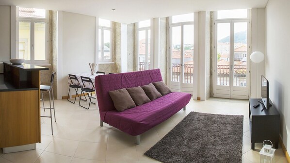 Una sala de estar moderna con un sofá púrpura, un comedor con mesa y sillas, y ventanas grandes que ofrecen vistas de la ciudad.