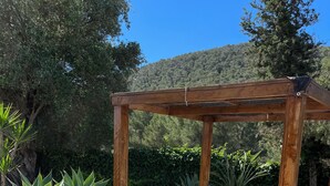 Een houten pergola met een matras en kussens, omringd door potplanten en een betegeld terras.