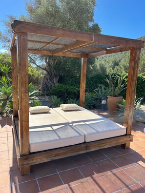 Une pergola en bois, agrémentée d’un matelas et de coussins, entourée de plantes et d’un arbre.