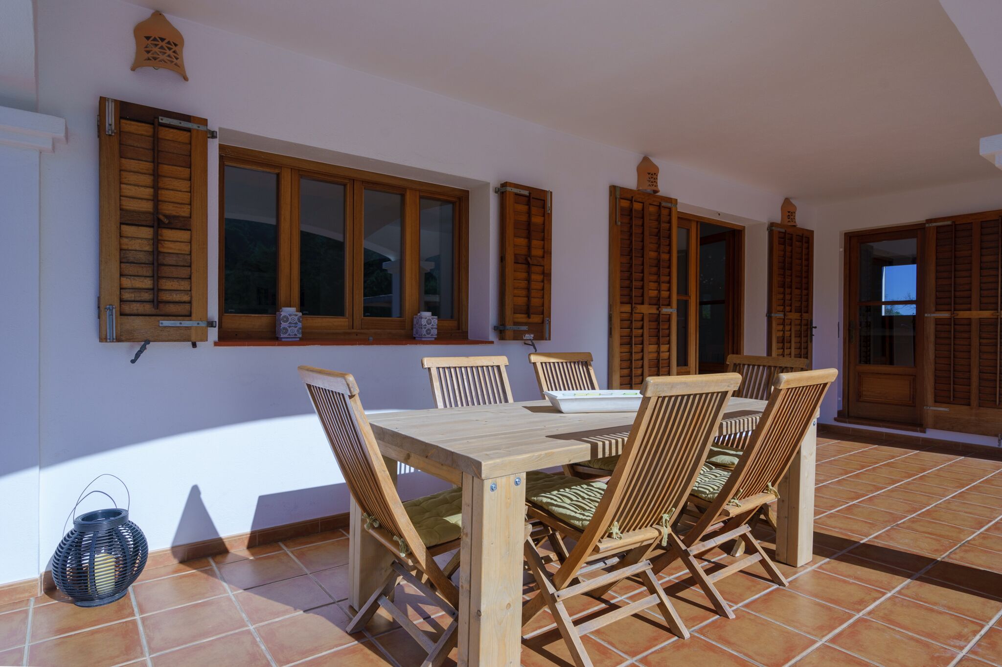 A dining area with a wooden table and chairs, large windows with wooden shutters, and a tiled floor.