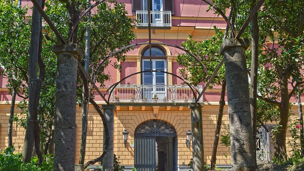 Un edificio con entrada roja, un balcón con barandilla blanca y una puerta con arco.