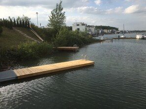 A wooden dock extending into a calm body of water, surrounded by greenery and buildings in the background. - Living right on the lake (Neukieritzsch)