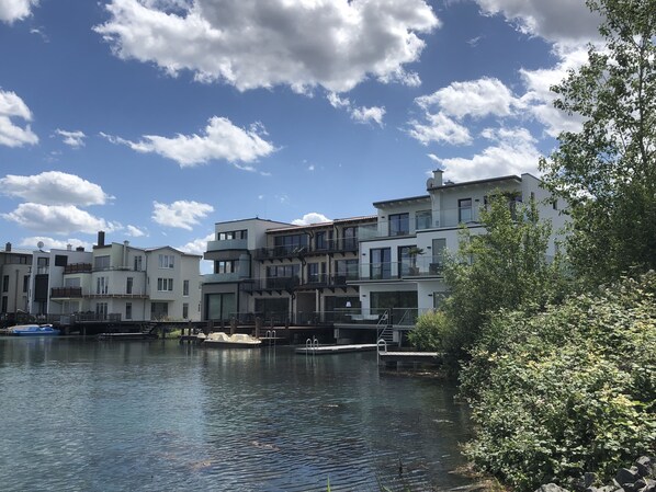 A waterfront residential area with modern multi-story buildings, a calm water body, and lush greenery. - Living right on the lake (Neukieritzsch)