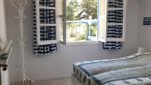Une chambre à coucher avec un lit, une fenêtre avec des rideaux, un petit tabouret et un porte-manteau fixé au mur.