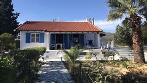 Une maison à deux étages avec un toit recouvert de tuiles rouges, des murs blancs et des volets bleus. On y trouve une petite terrasse avec une table et des chaises, ainsi qu’un escalier menant à l’étage supérieur.