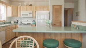 A kitchen with wooden cabinets, a white refrigerator, a microwave, a stove, and a green countertop. There are two green stools and a chair. - Lovely 4 Bedroom Ocean view house in West Beach (Oak Island)