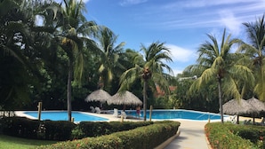 A pool area with thatched umbrellas, palm trees, and a clear blue sky. - Luxury And Peaceful Little Condo With Direct Access To The Beach (Zihuatanejo)