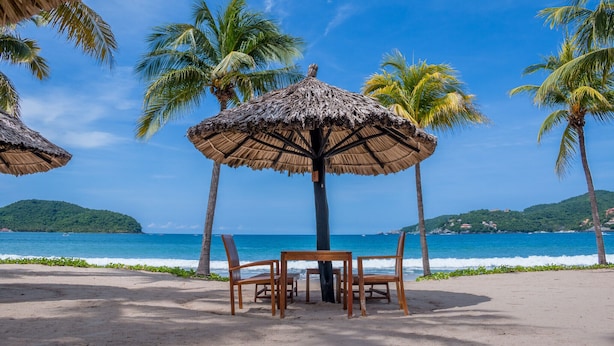 A beachside setting with a table and chairs under a thatched umbrella, surrounded by palm trees and a view of the ocean. - Luxury And Peaceful Little Condo With Direct Access To The Beach (Zihuatanejo)