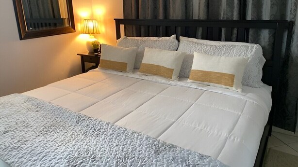 Una cama bien hecha con sábanas blancas y una manta con textura, una lámpara de noche y un espejo en la pared.