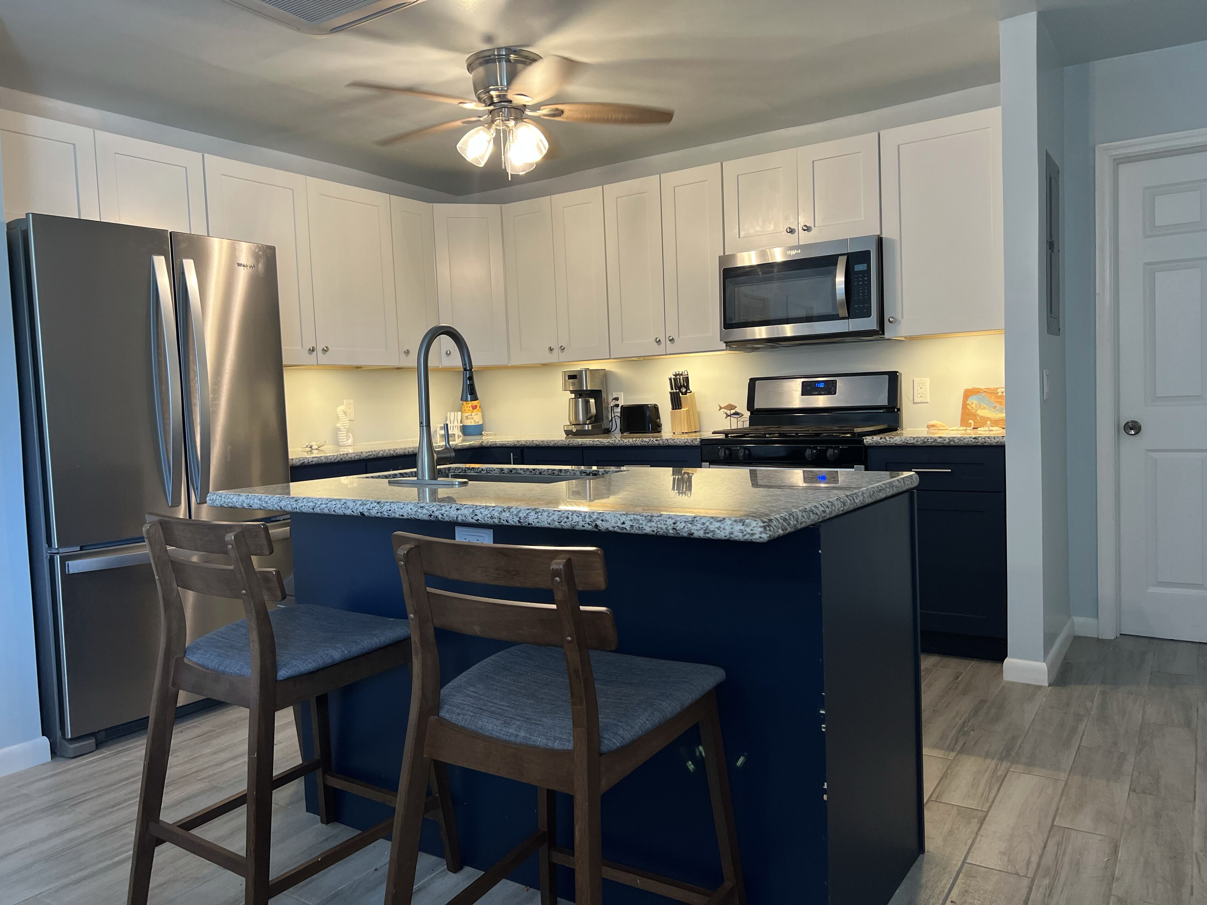 A modern kitchen with white cabinets, a stainless steel refrigerator, a microwave, a stove, and a ceiling fan. There are two wooden bar stools with blue cushions.