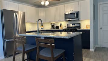 A modern kitchen with white cabinets, a stainless steel refrigerator, a microwave, a stove, and a ceiling fan. There are two wooden bar stools with blue cushions.