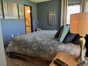 Un dormitorio con una colcha estampada, una guitarra, un tapiz de macramé y una lámpara en una mesita de noche.