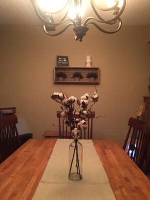 Una mesa de comedor con un jarrón de flores secas, sillas de madera, una araña, una estantería con plantas y un cartel enmarcado encima de la estantería.