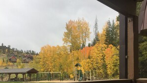 Una terraza de madera con sillas de mimbre y una mesa, con vista a un parque infantil y árboles de colores otoñales.