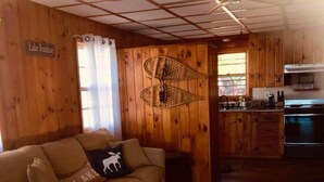 A cozy cabin interior with a wooden floor, a sofa with a pillow, a kitchen area with a stove, and a window with curtains. - Sandy Beach Oasis on Lake Ivanhoe (Wakefield)