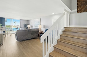 Un vaste espace de vie comprenant un escalier, un canapé gris et une vue sur l’extérieur.